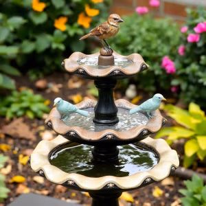 En brun fågel sitter på toppen av det tvådelade fågelbadet fågelbad.se, med dekorativa blå fågelfigurer och omgivet av frodig grönska med blommande rosa och gula blommor.