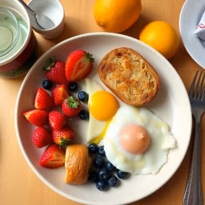 På en tallrik från brunchgöteborg.se finns två solvända ägg, rostat bröd, jordgubbar, blåbär och en bulle. I närheten finns citroner, en kopp, en gaffel, en mjölkkanna och ett inslaget föremål.
