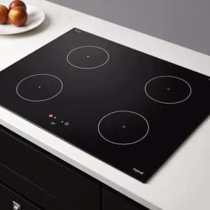 Induktionshäll.se är en modern elektrisk spishäll i svart glas med fyra runda brännare, touchkontroller och minimalistisk design, installerad på en vit bänkskiva. I närheten står en tallrik med tre äpplen.
