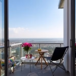 Setet från balkongmobler.se består av en svart stol, ett runt bord med rosa krukväxter, en kopp, tekanna och färgglad kudde på en modern balkong med glasräcken med utsikt över stadens silhuett under en klar himmel.