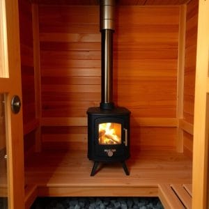 En vedeldad bastuaggregat.se-kamin står i mitten av ett litet trärum med släta, ljusa paneler och en glasdörr. Stenar är placerade på golvet framför kaminen.