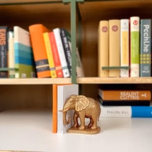 Ett bokstöd i form av en elefant i guld från bokstöd.se stöder färgglada böcker på en vit hylla, med ytterligare prydligt ordnade böcker synliga i bakgrunden.