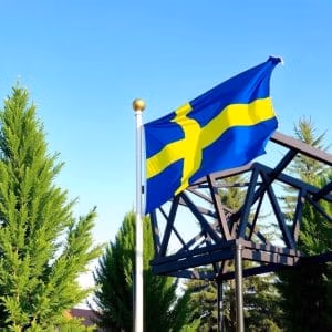 Den svenska flaggan flaggstang.se, med ett gult nordiskt kors på blå botten, vajar utomhus på en flaggstång bland gröna träd och en metallkonstruktion under en klar himmel.