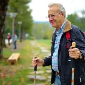En leende äldre man med grått hår och glasögon håller i gåstavar.se vandringsstavar, klädd i rutig skjorta, svart jacka och ryggsäck när han står på en parkväg omgiven av träd och en träbänk.