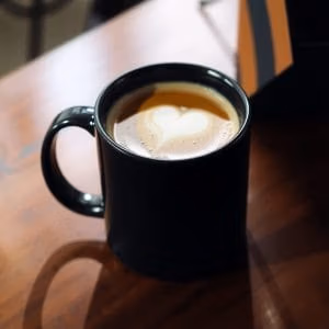 En kaffemugg.se i svart keramik fylld med kaffe, med hjärtformad skumdesign, står på ett träbord i mjukt dagsljus.