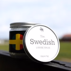 En metallburk märkt "lössnus.se" med en kamelikon vilar på en avsats utomhus. På sidan av burken syns en design inspirerad av den svenska flaggan, med suddigt gräs och himmel i bakgrunden.