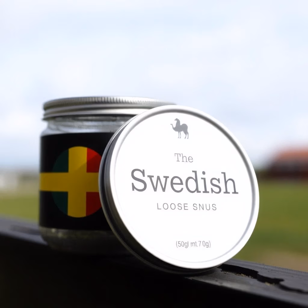 En metallburk märkt "lössnus.se" med en kamelikon vilar på en avsats utomhus. På sidan av burken syns en design inspirerad av den svenska flaggan, med suddigt gräs och himmel i bakgrunden.