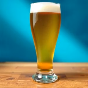 Ett högt glas från ölglas.se, fyllt med gyllene öl och toppat med ett vitt skumhuvud, står på ett träbord mot en livligt blå bakgrund.