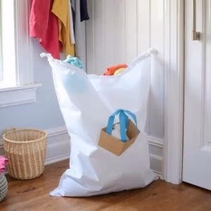 En tvättsäck från tvättpåse.se, fylld med färgglada kläder, vilar på ett trägolv bredvid en vit vägg och dörr. En liten korg och hängande kläder syns också i bakgrunden.